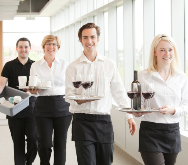 A group of four smiling hospitality staff members are walking down a bright hallway. They are dressed in white shirts and black aprons. The staff members are carrying trays with wine glasses, a wine bottle, and coffee cups, showcasing a professional and welcoming service environment.