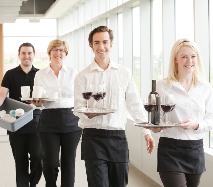 A group of four smiling hospitality staff members are walking down a bright hallway. They are dressed in white shirts and black aprons. The staff members are carrying trays with wine glasses, a wine bottle, and coffee cups, showcasing a professional and welcoming service environment.