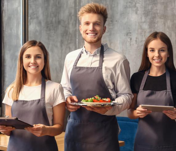 Three smiling hospitality workers in aprons stand side by side in a restaurant setting. The woman on the left holds a clipboard, the man in the middle holds a plate of food, and the woman on the right holds a tablet, showcasing teamwork and professionalism in the hospitality industry.