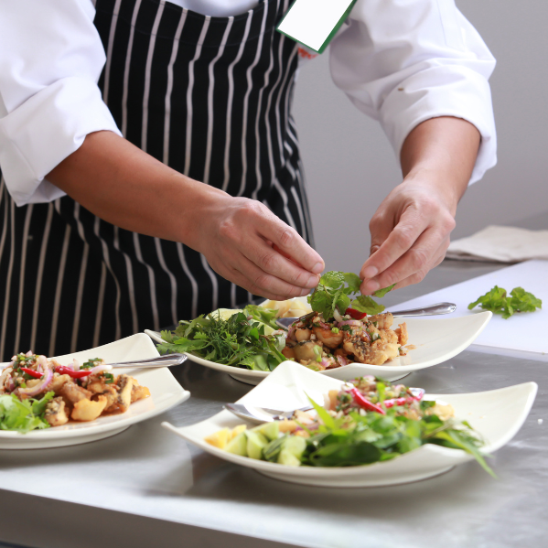 A chef wearing a white uniform and a black-and-white striped apron is carefully garnishing plates of food with fresh herbs. The dishes are elegantly arranged with a variety of colourful ingredients, and the scene takes place in a professional kitchen setting.