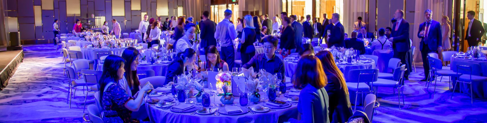 The President's Dinner & Training Awards. A vibrant evening event in a grand ballroom filled with elegantly set round tables. Guests are seated and socializing, with some standing and mingling in the background. The tables are adorned with formal table settings, including glassware, folded napkins, and decorative centerpieces, all under a soft, bluish lighting that gives the room a luxurious ambiance.