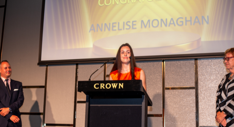 A young woman, Annelise Monaghan, stands at a podium marked 'CROWN,' delivering a speech at an awards ceremony. She is flanked by two individuals, one on each side, as she accepts an award. The screen behind her displays her name and the word 'Congratulations.' The setting is formal and celebratory.