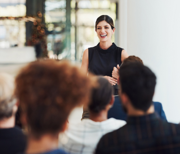 A woman in a sleeveless black top stands smiling in front of a group of seated people, engaging them in a discussion or presentation. The setting appears to be a modern, bright conference or training room, emphasizing communication and leadership.